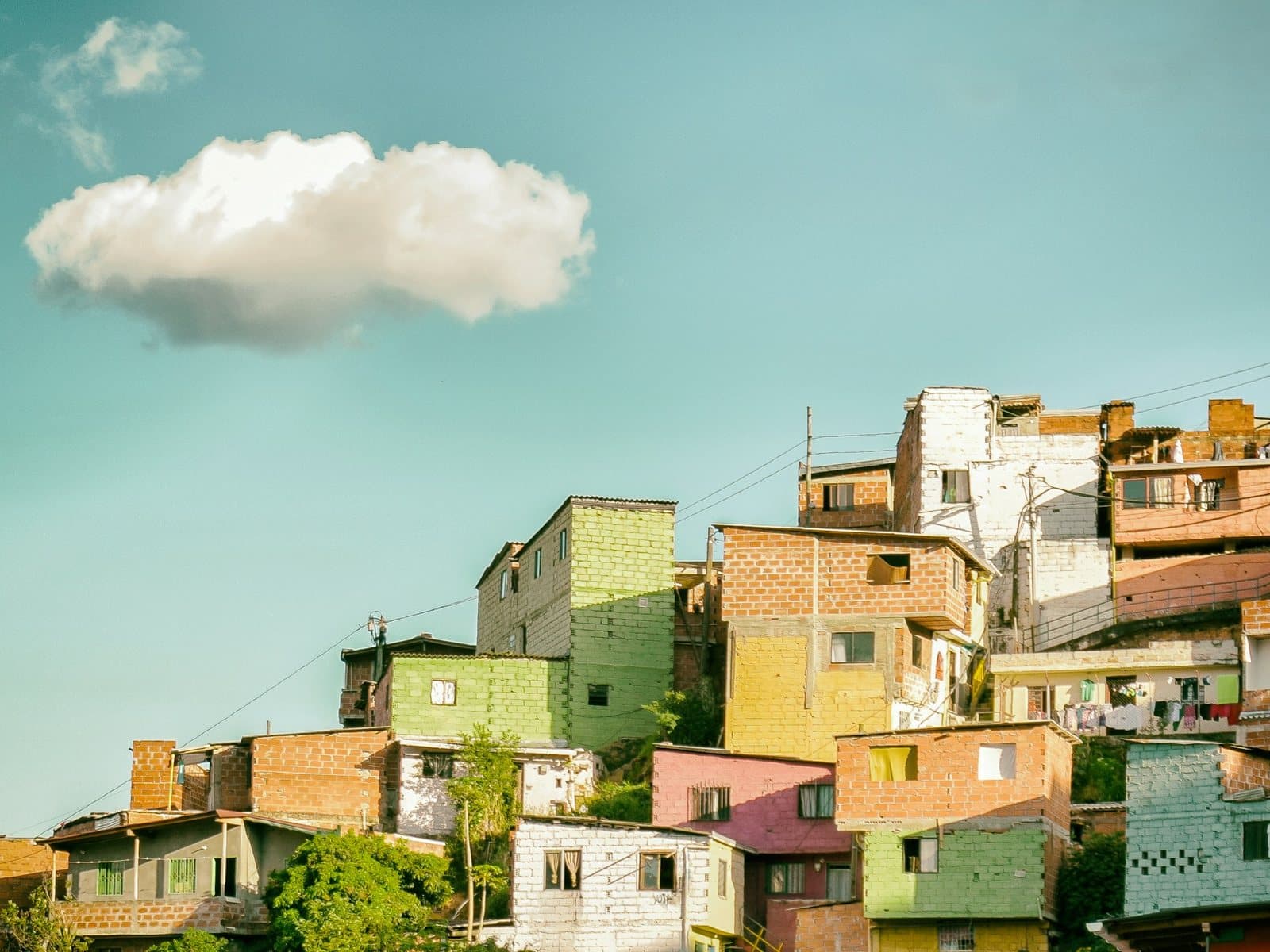 Pastel hillside homes in yellow, green and pink stack up across Comuna 13 under teal sky