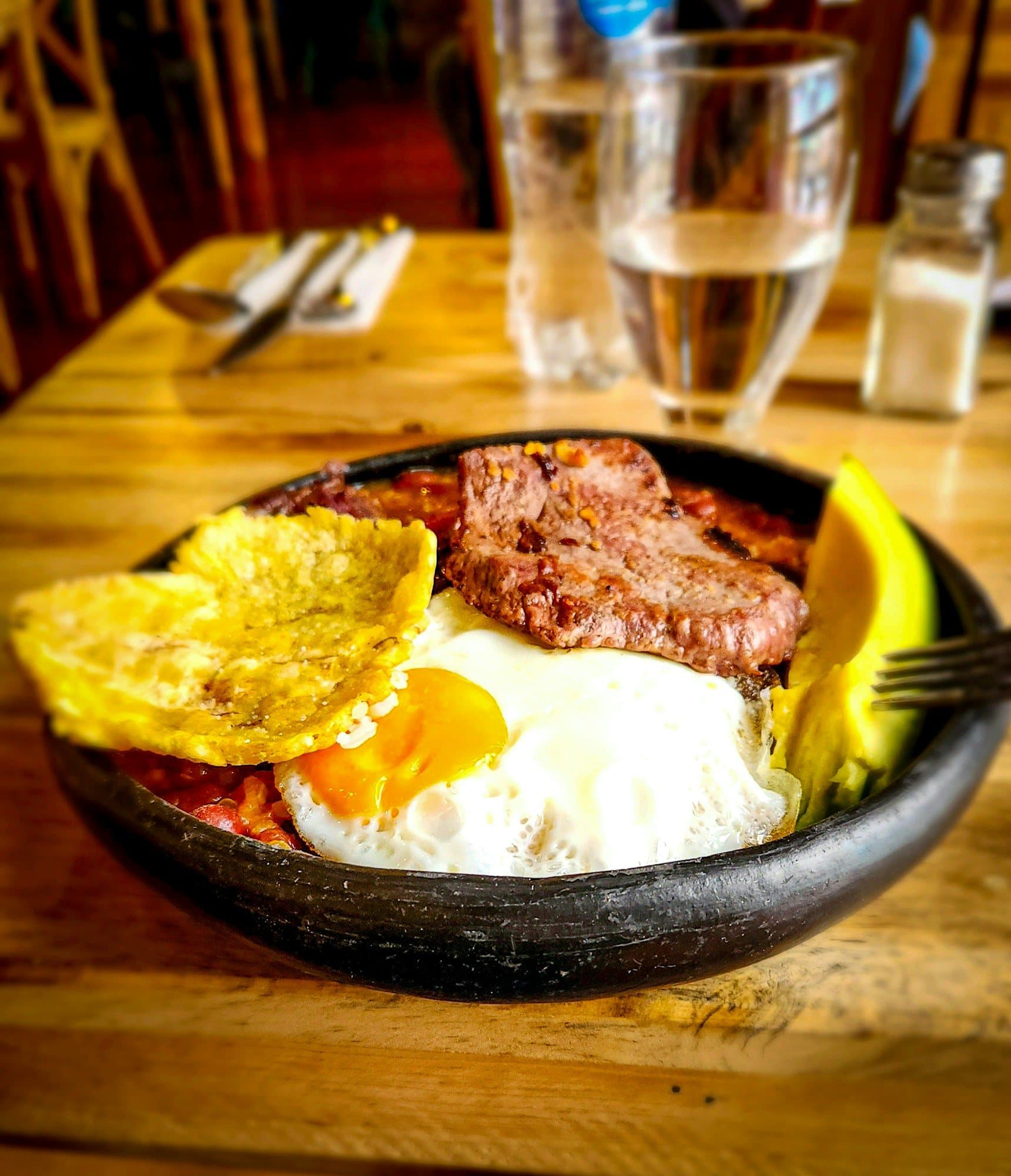 Una cazuela de barro chisporroteante con desayuno colombiano: huevo frito, carne, patacón crujiente y aguacate