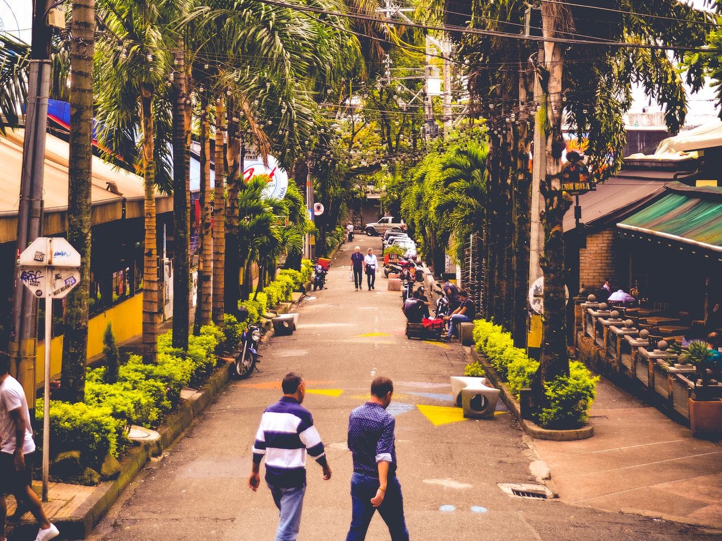 Gente caminando por una calle frondosa con palmeras, plantas y terrazas de cafés en El Poblado