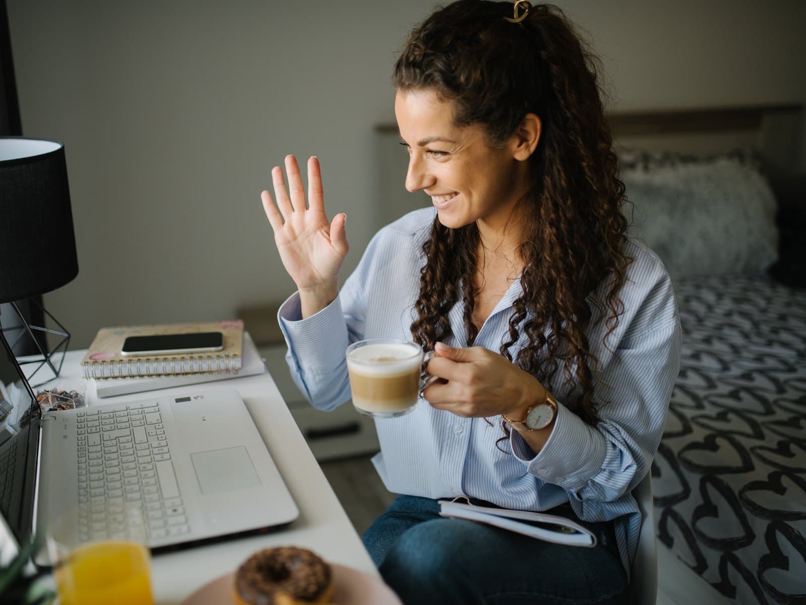 Smiling woman with coffee waving warmly during a friendly video call at her laptop