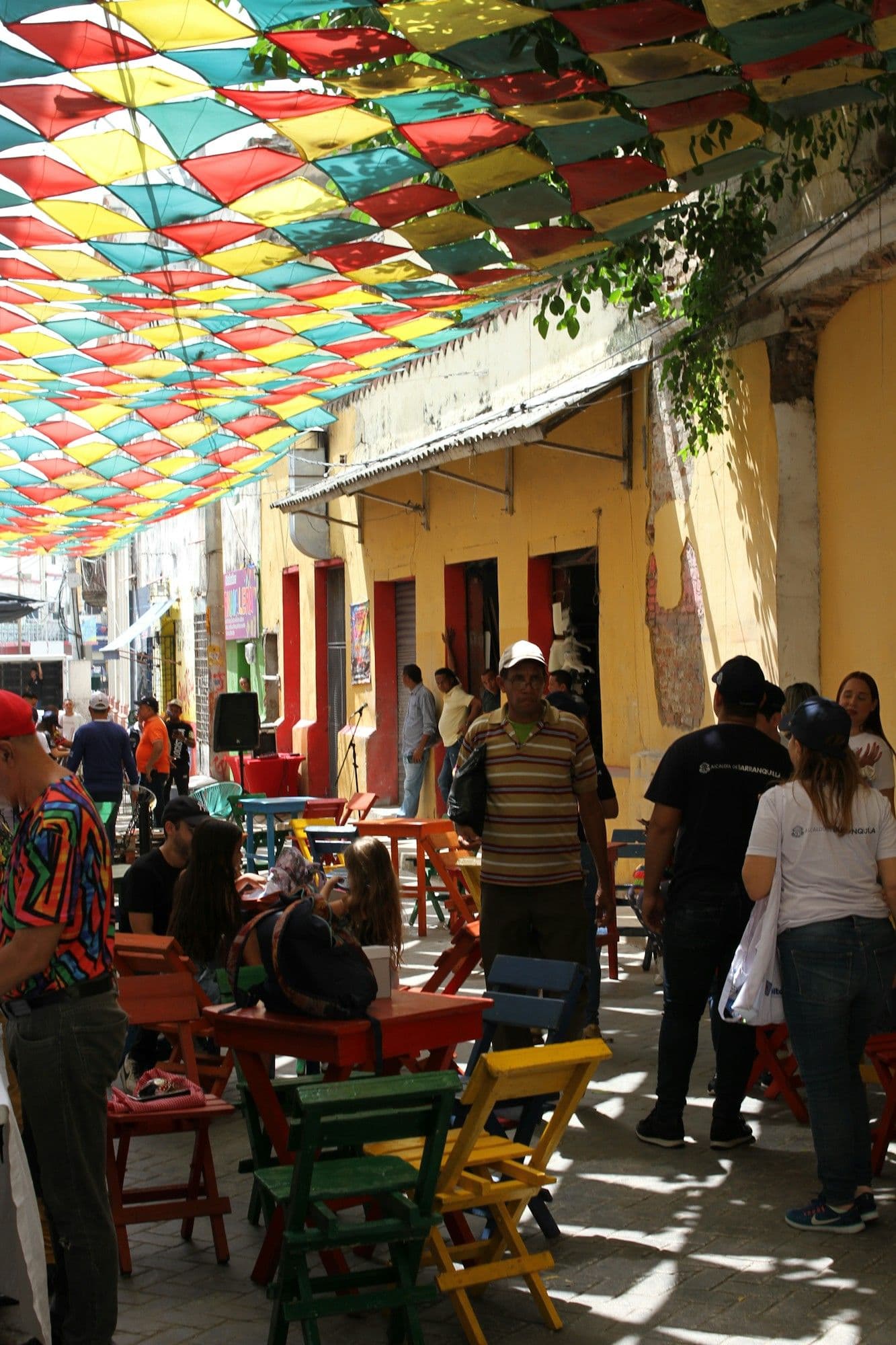 Friends gather at colorful tables beneath a rainbow canopy of pennants on a sunny street