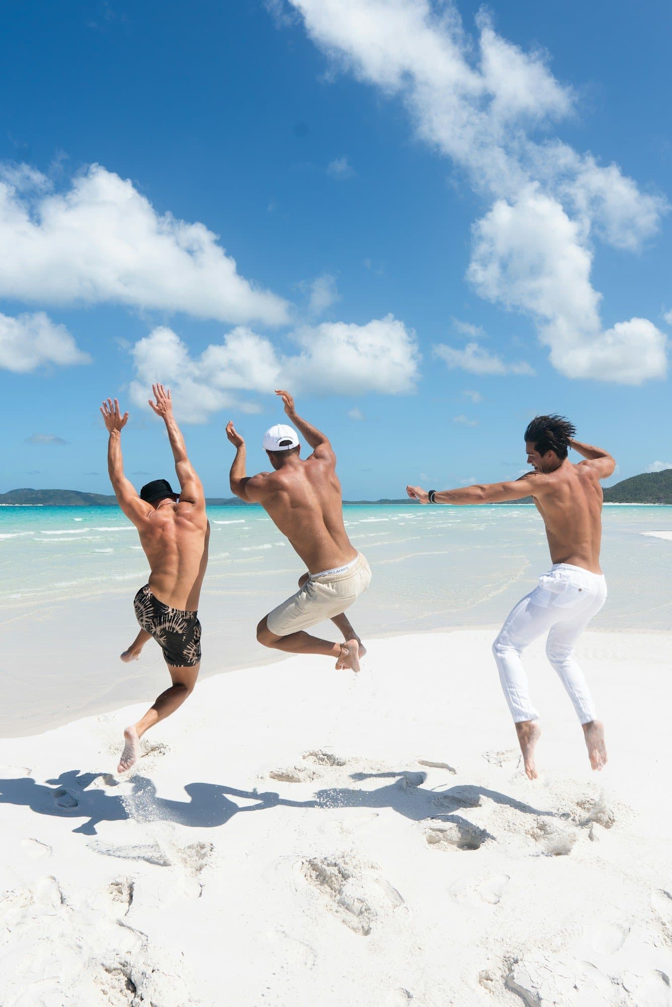 Three friends leap for joy on a white-sand beach under a bright blue sky