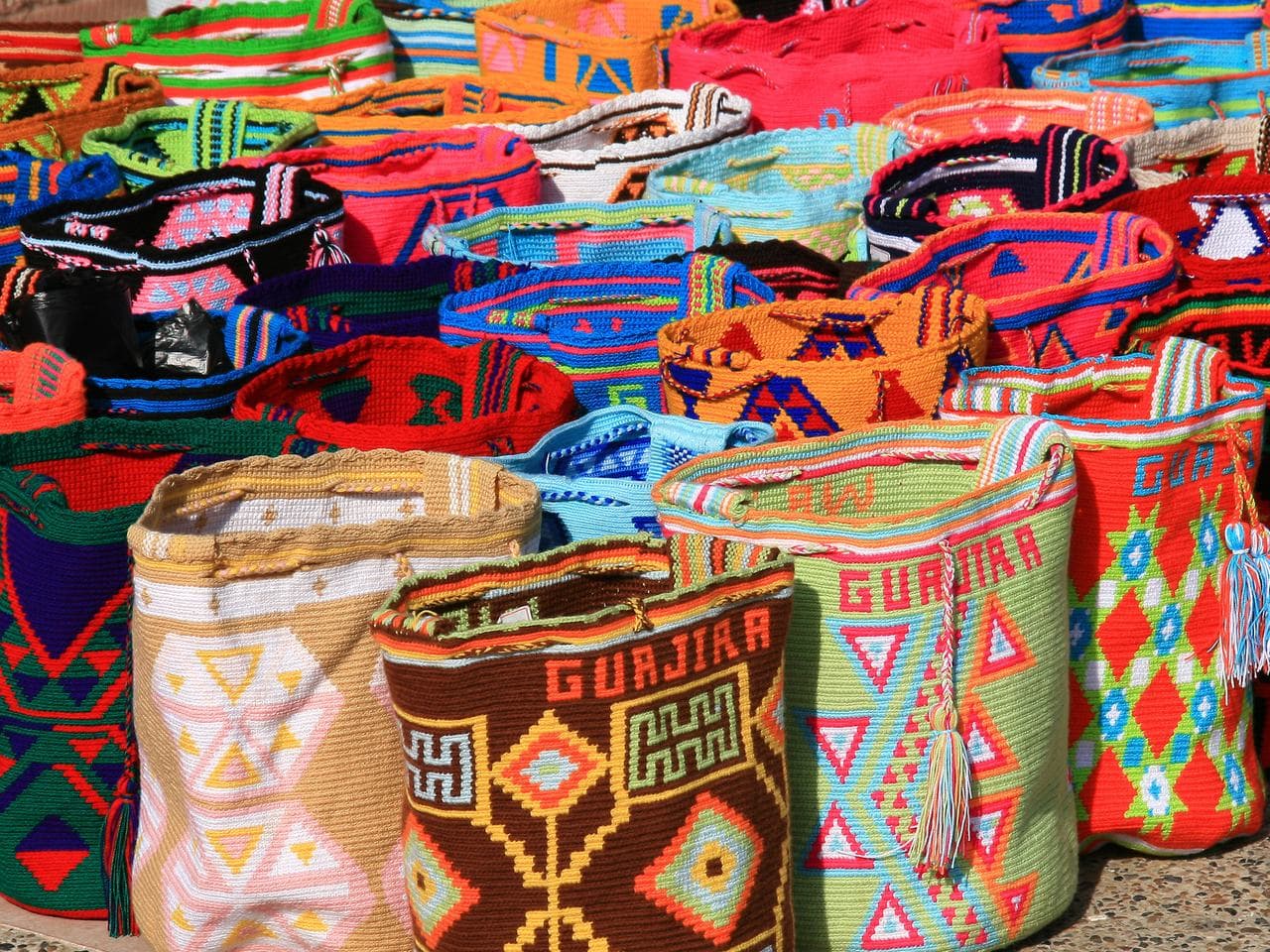 Stacks of handwoven Wayuu mochila bags in vivid geometric patterns piled at a market stall
