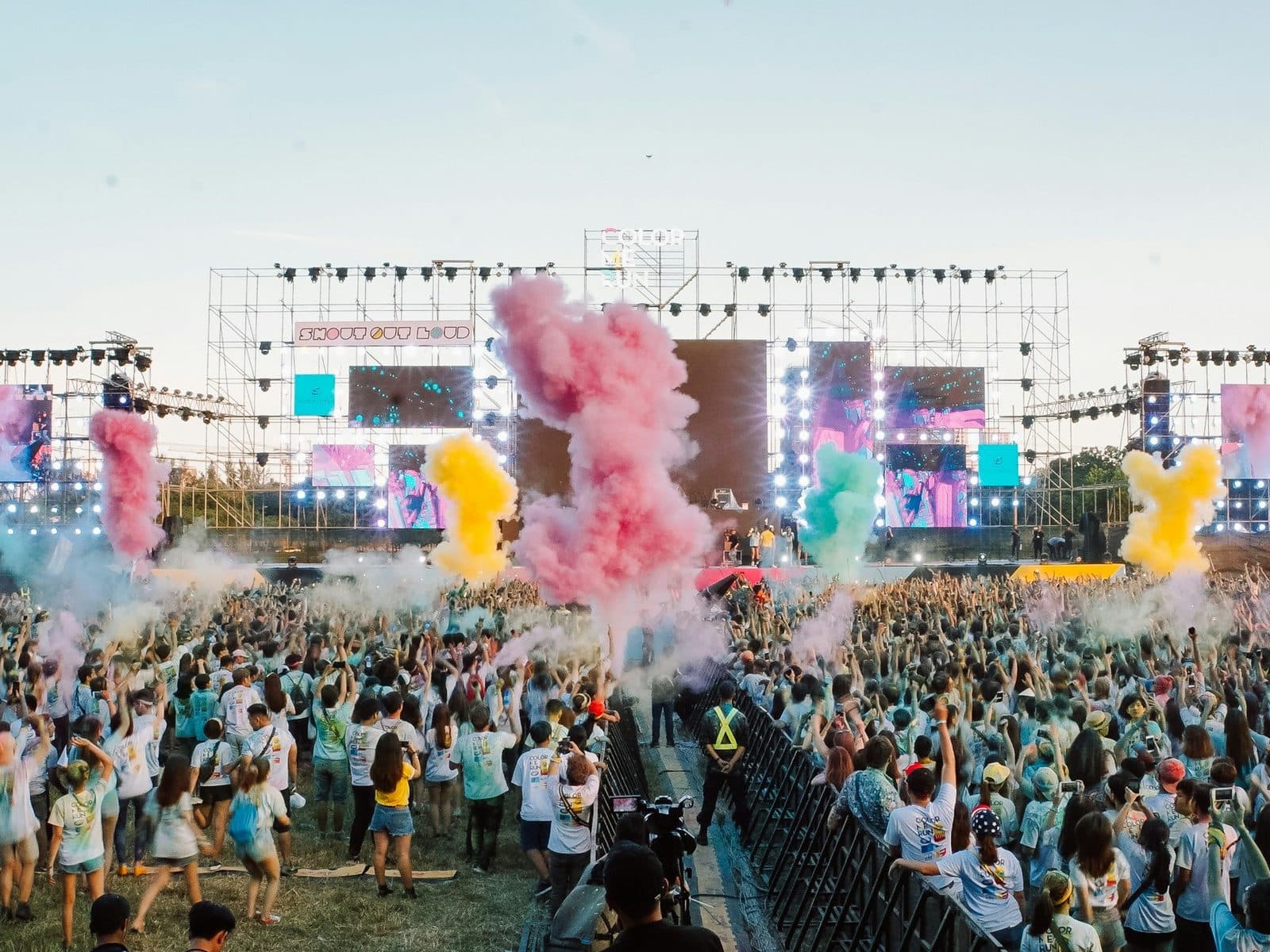 A huge open-air festival crowd dances as pink, yellow and teal smoke rises before the stage