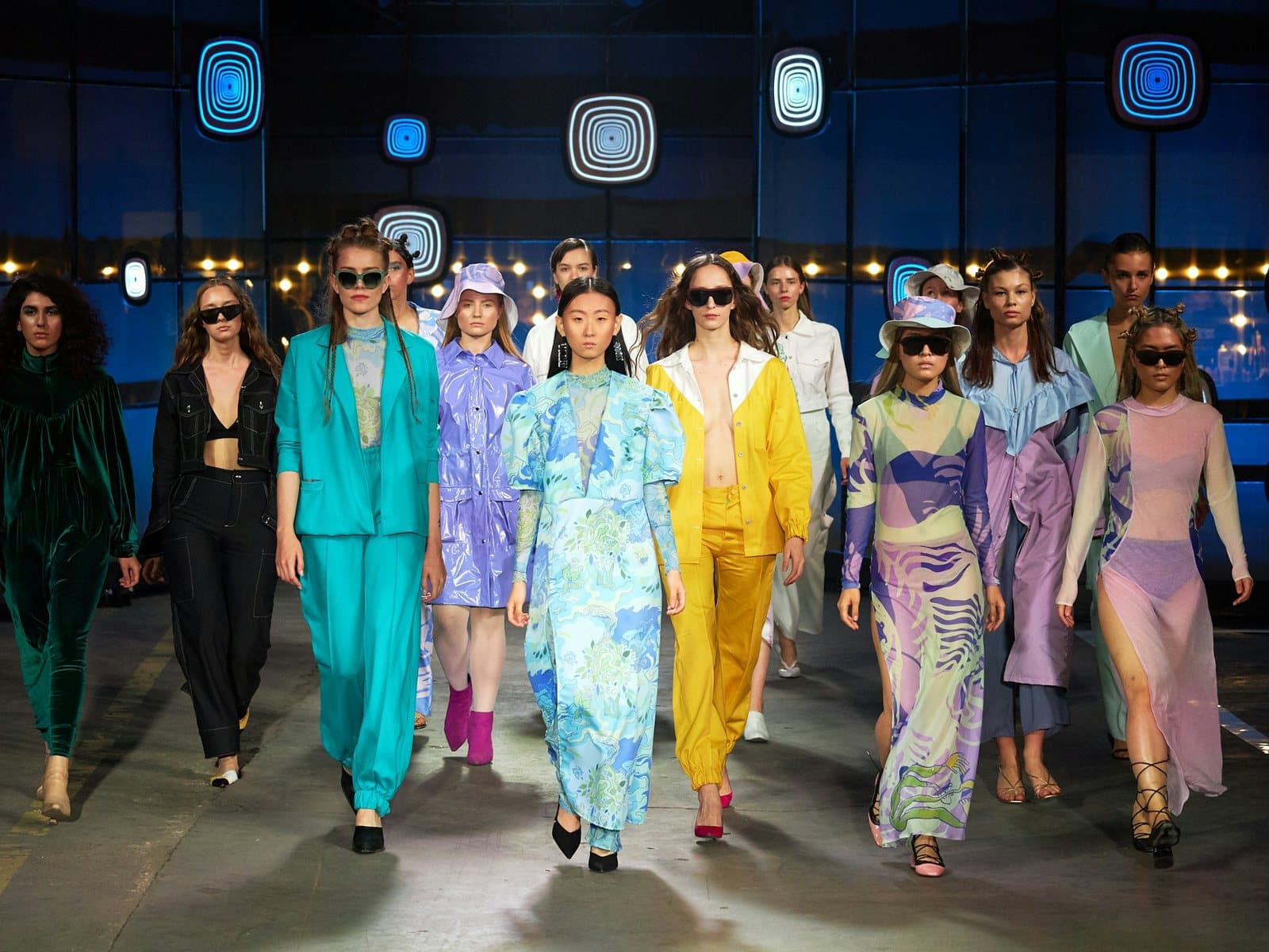 Models in turquoise, yellow and lilac outfits walk the runway together at a fashion show finale