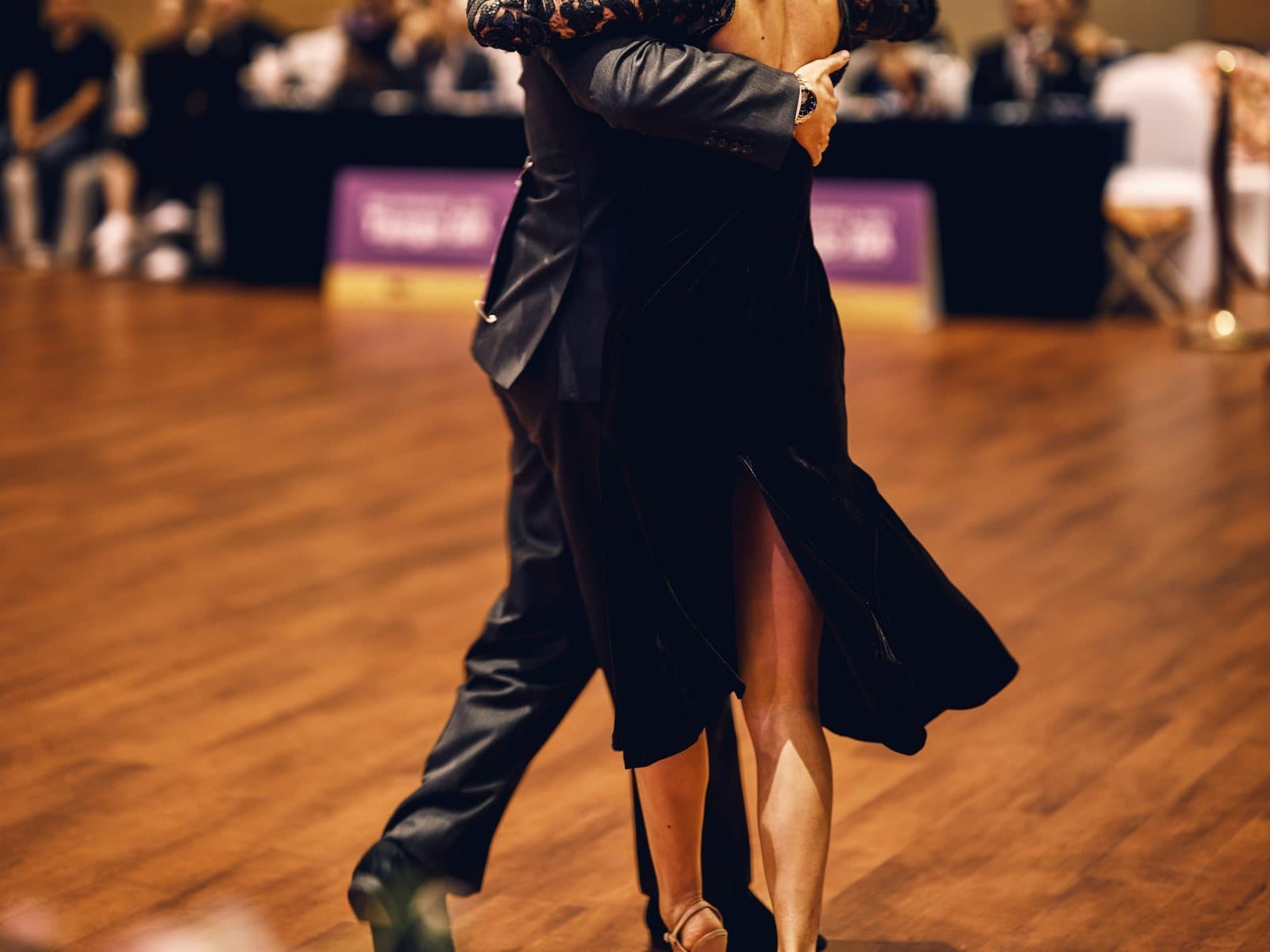 A tango couple in elegant black glides across a warm wooden dance floor at a milonga