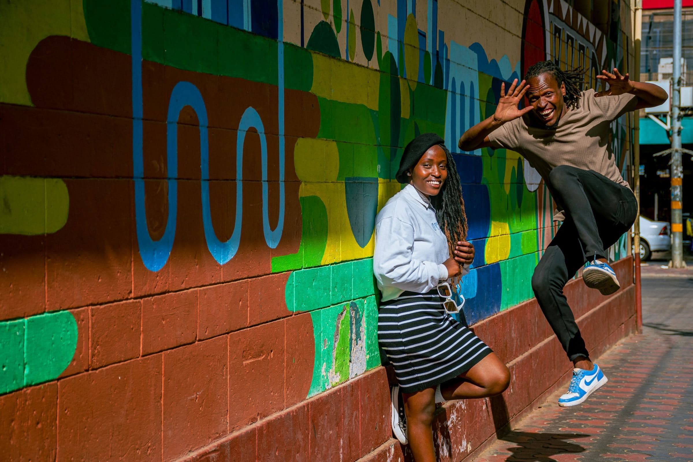 Two friends laugh and leap in front of a bold green, blue and red mural