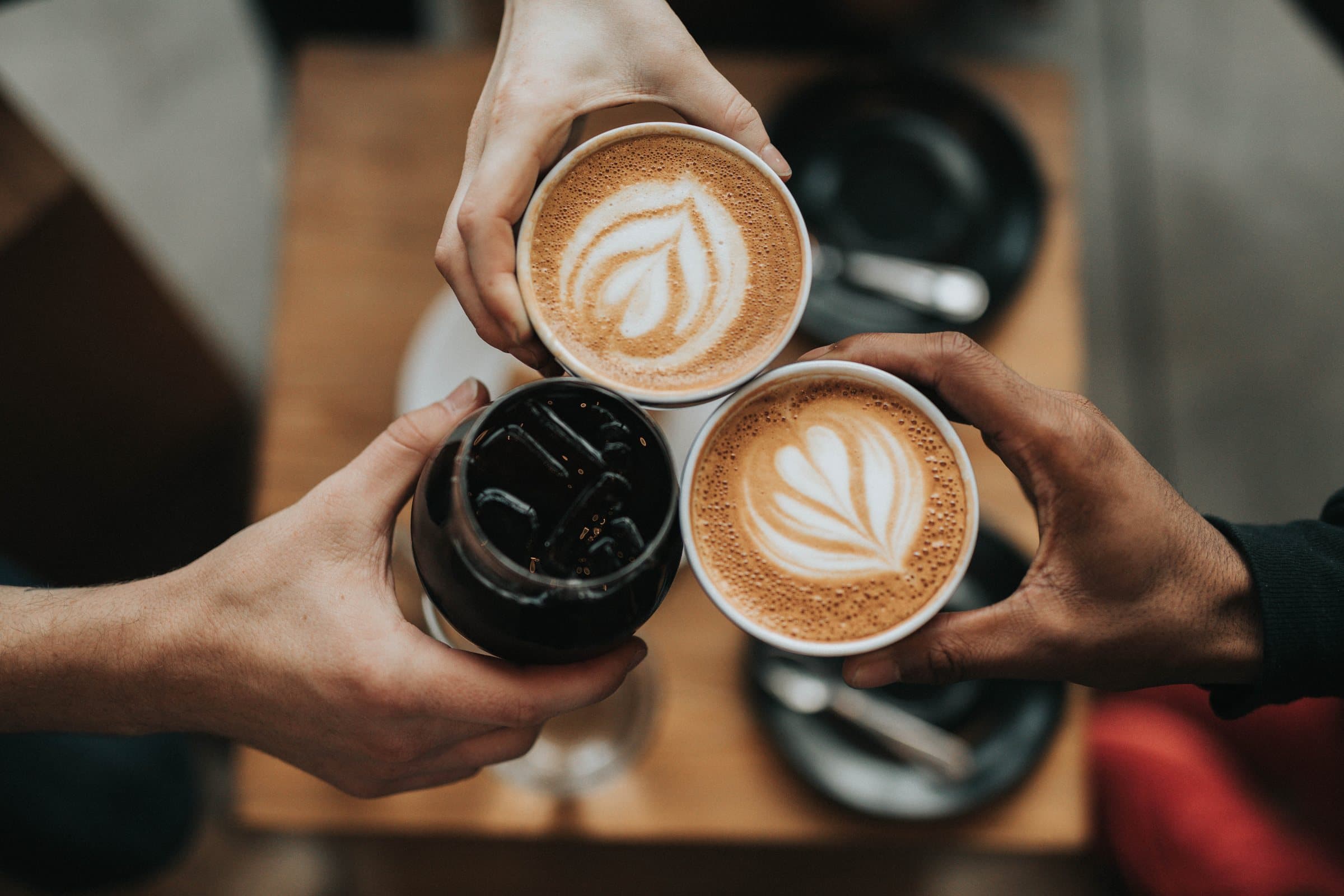 Three friends clink latte-art coffees and an iced brew together over a café table