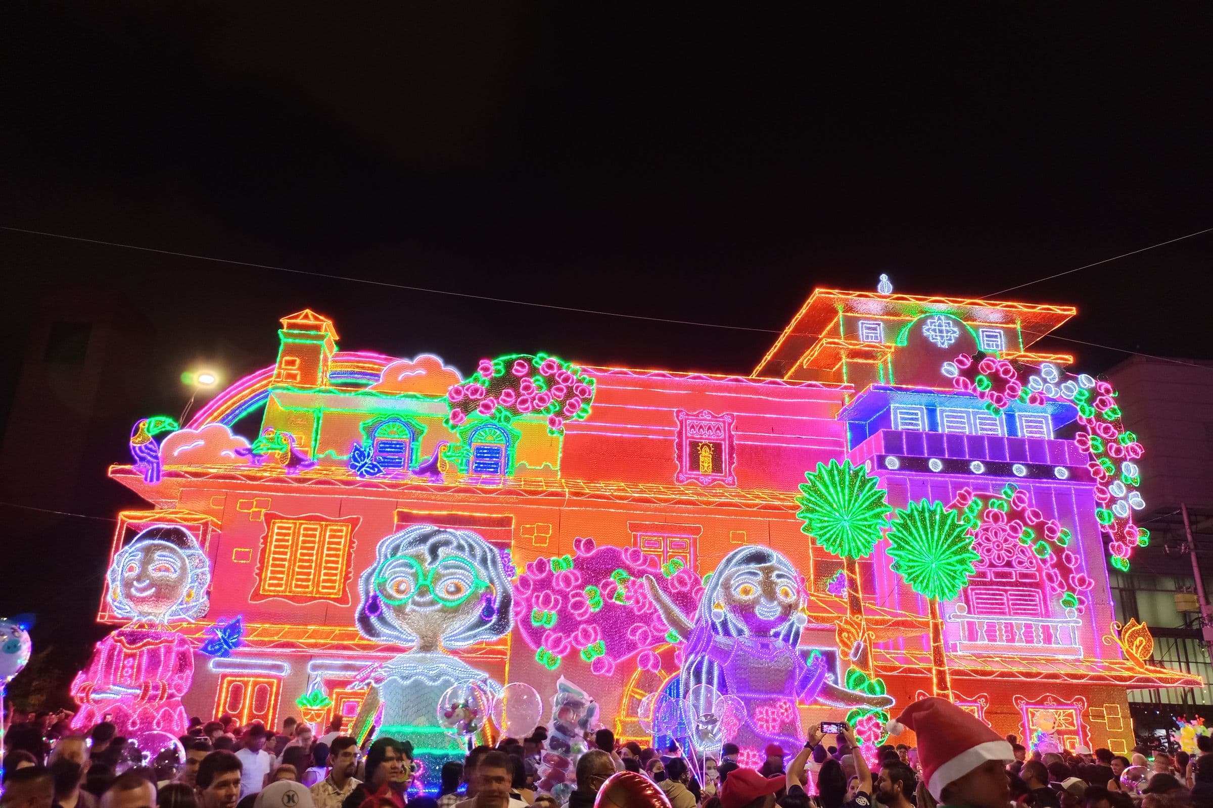 Neon Christmas light display of a colorful house glows above crowds at Medellín's Alumbrados