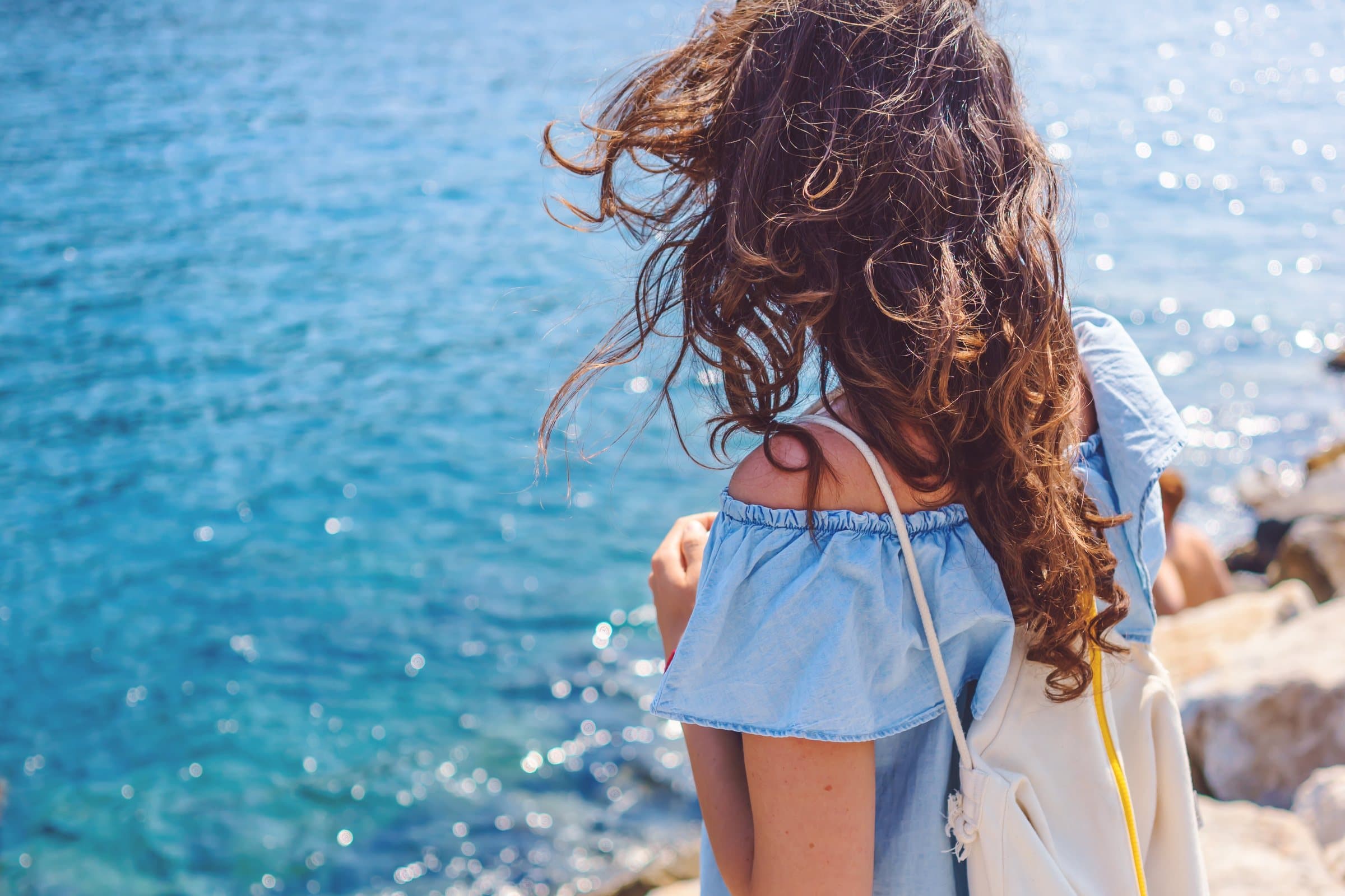 Viajera con mochila y rizos al viento contempla un mar azul resplandeciente
