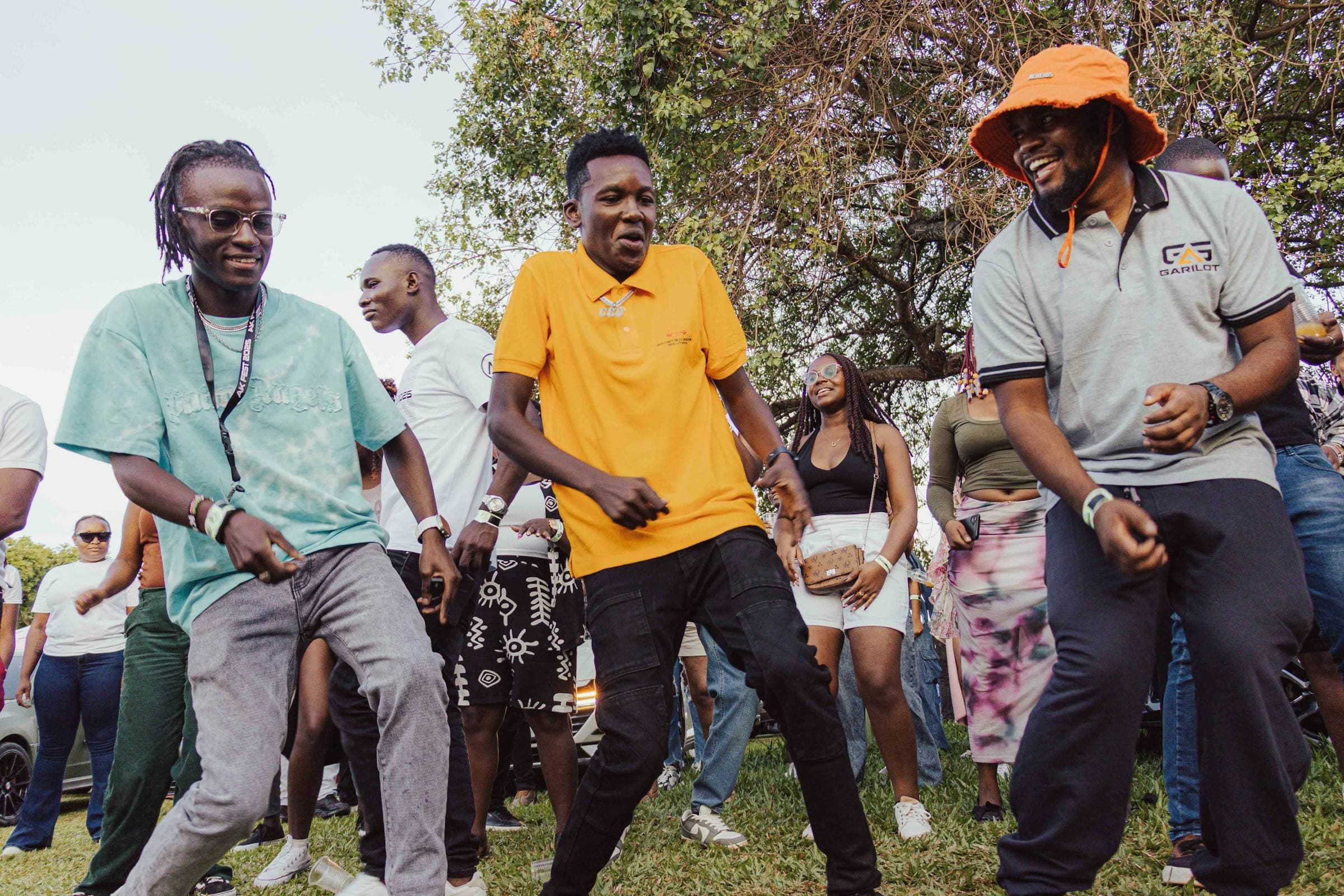 Friends dance and laugh together at a sunny outdoor party, one in a bright orange bucket hat