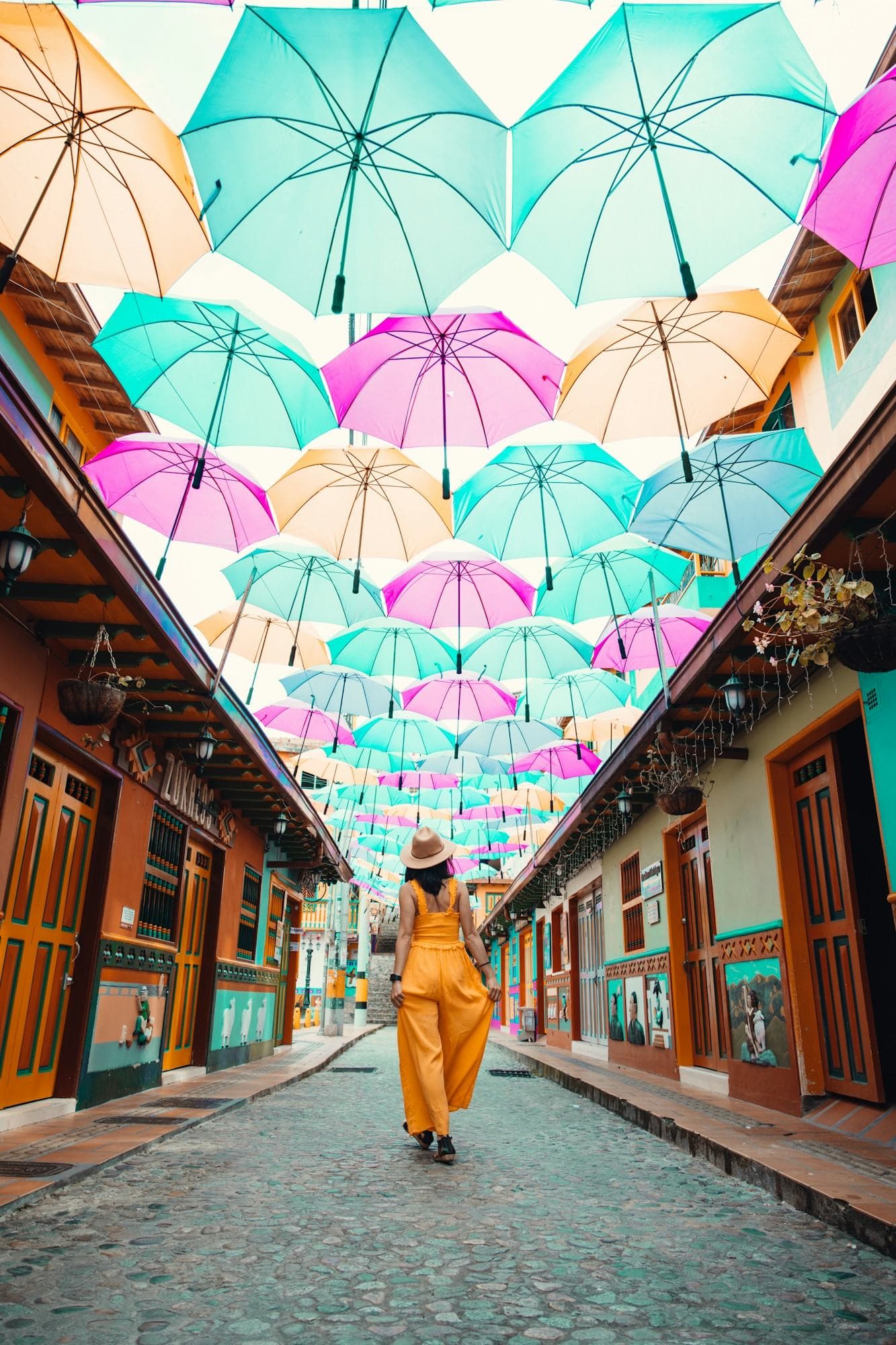 Una viajera con abrigo amarillo camina bajo un techo de sombrillas pastel en una colorida calle de Guatapé