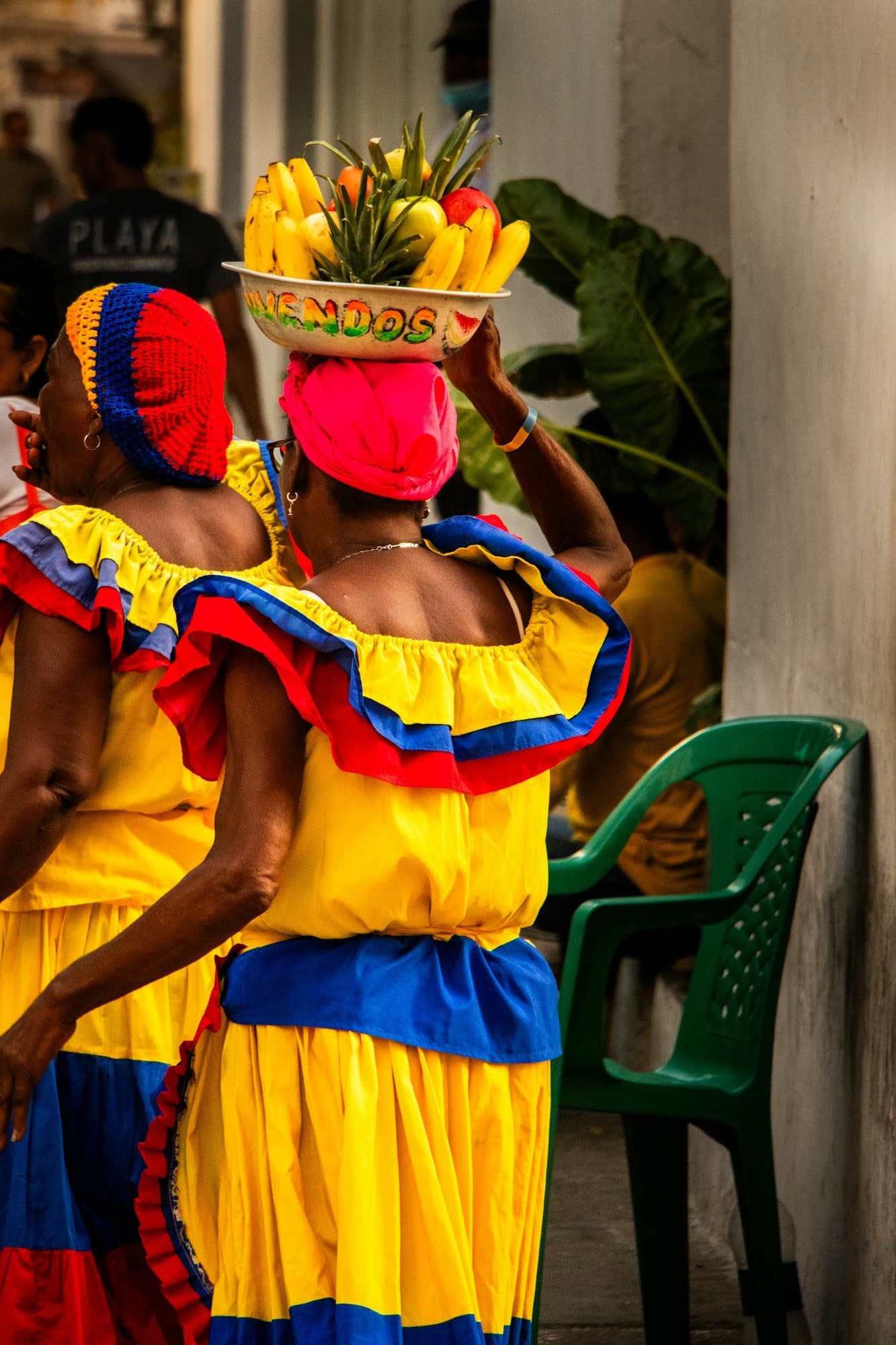 Dos palenqueras con vestidos amarillos, azules y rojos equilibran platones de fruta tropical en la cabeza en Cartagena
