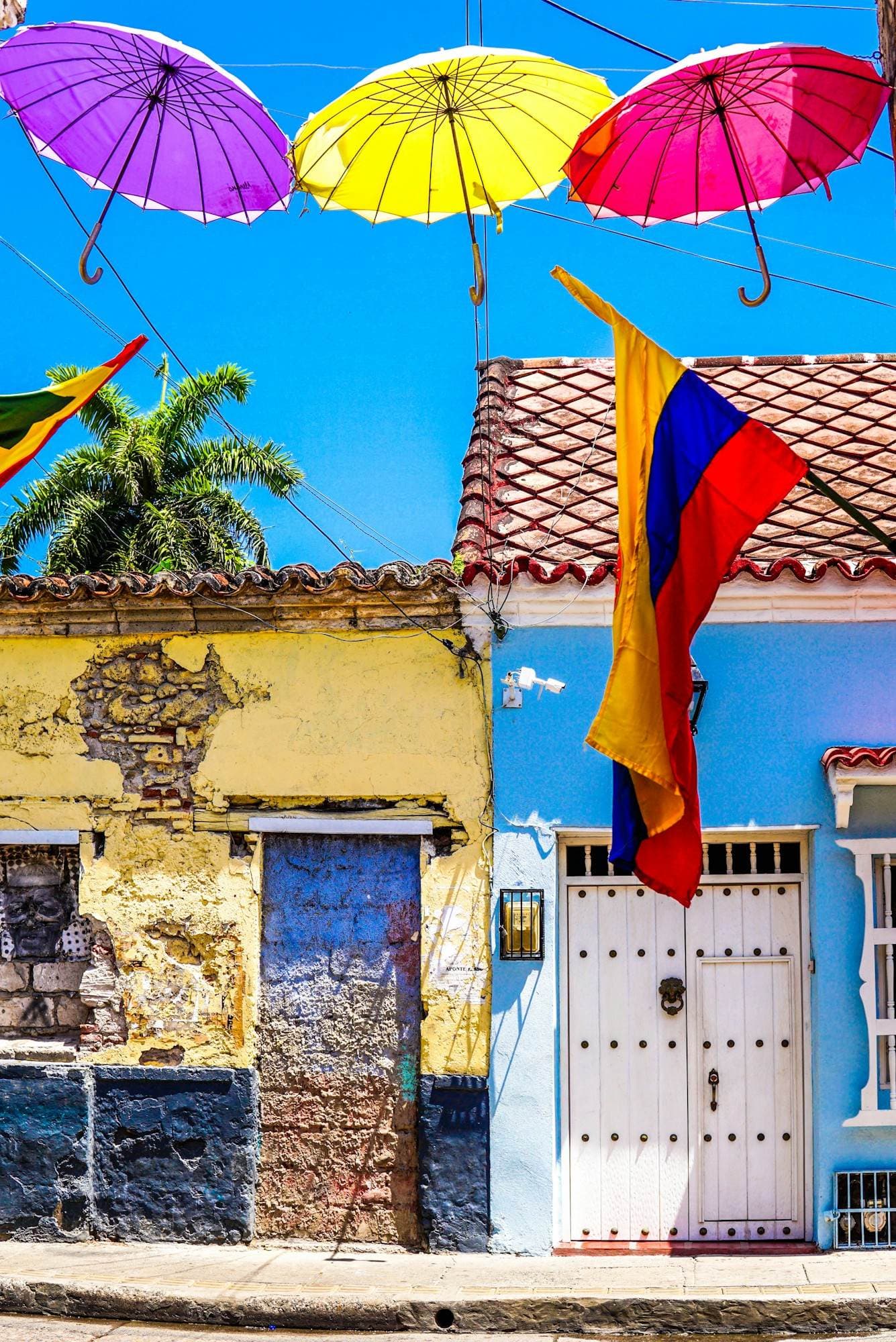 Sombrillas de colores flotan sobre una fachada vibrante y una bandera de Colombia ondeando en la calle de las sombrillas de Cartagena