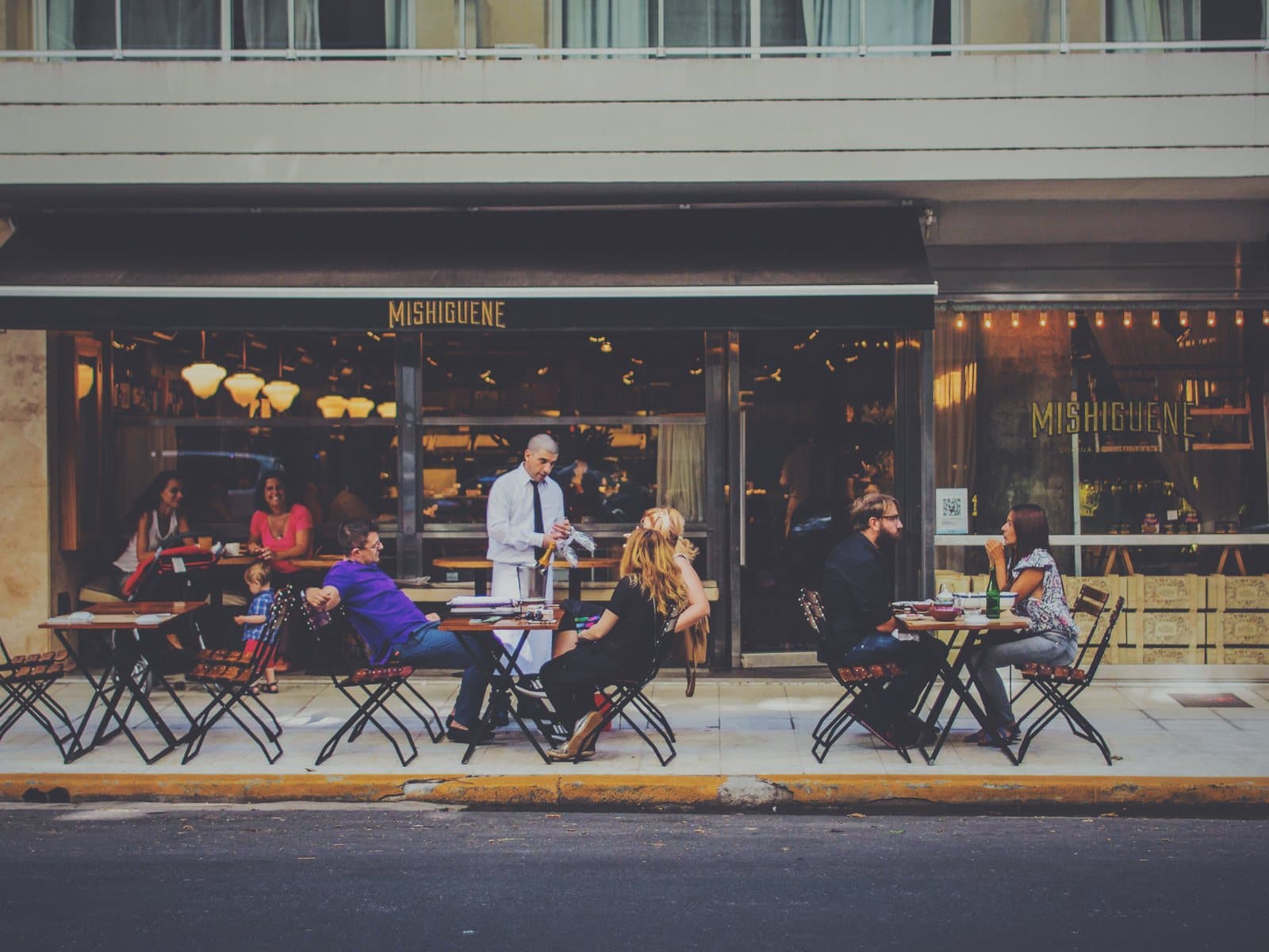Amigos conversan en mesas de un café en la acera bajo luces cálidas en una tarde soleada