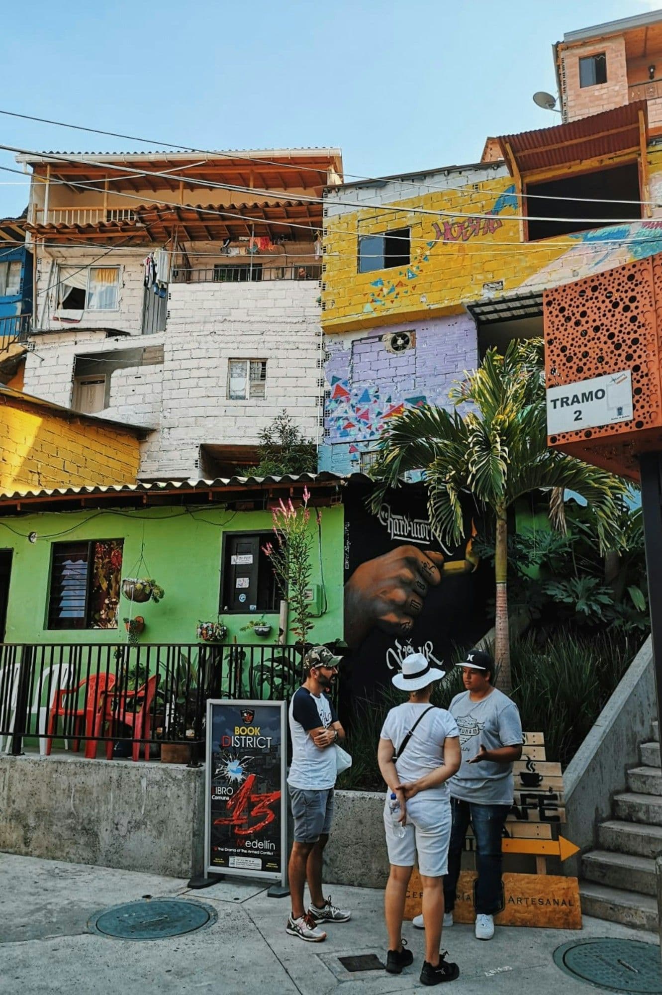 Viajeros conversan en una esquina soleada de la Comuna 13 bajo casas apiladas pintadas de amarillo, verde y rosado