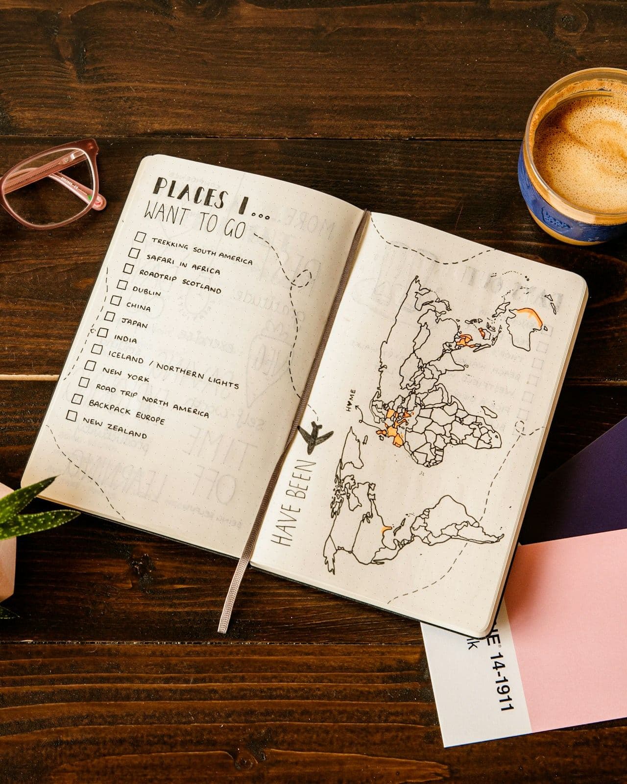 Un diario de viaje con una lista de lugares por visitar, abierto junto a un café recién hecho, unas gafas y una suculenta