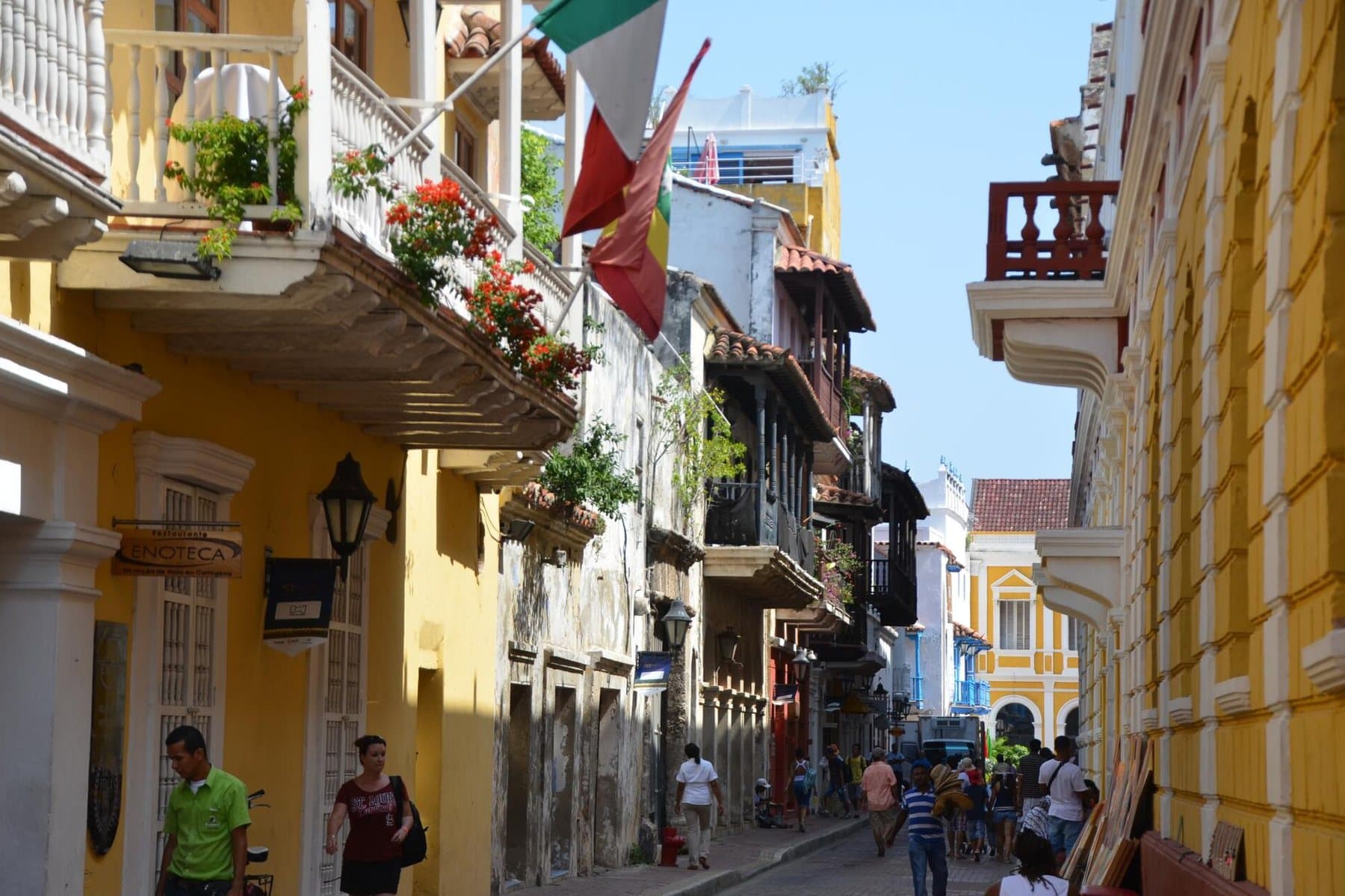 Calle colonial soleada con fachadas amarillas, balcones con flores, banderas y gente paseando