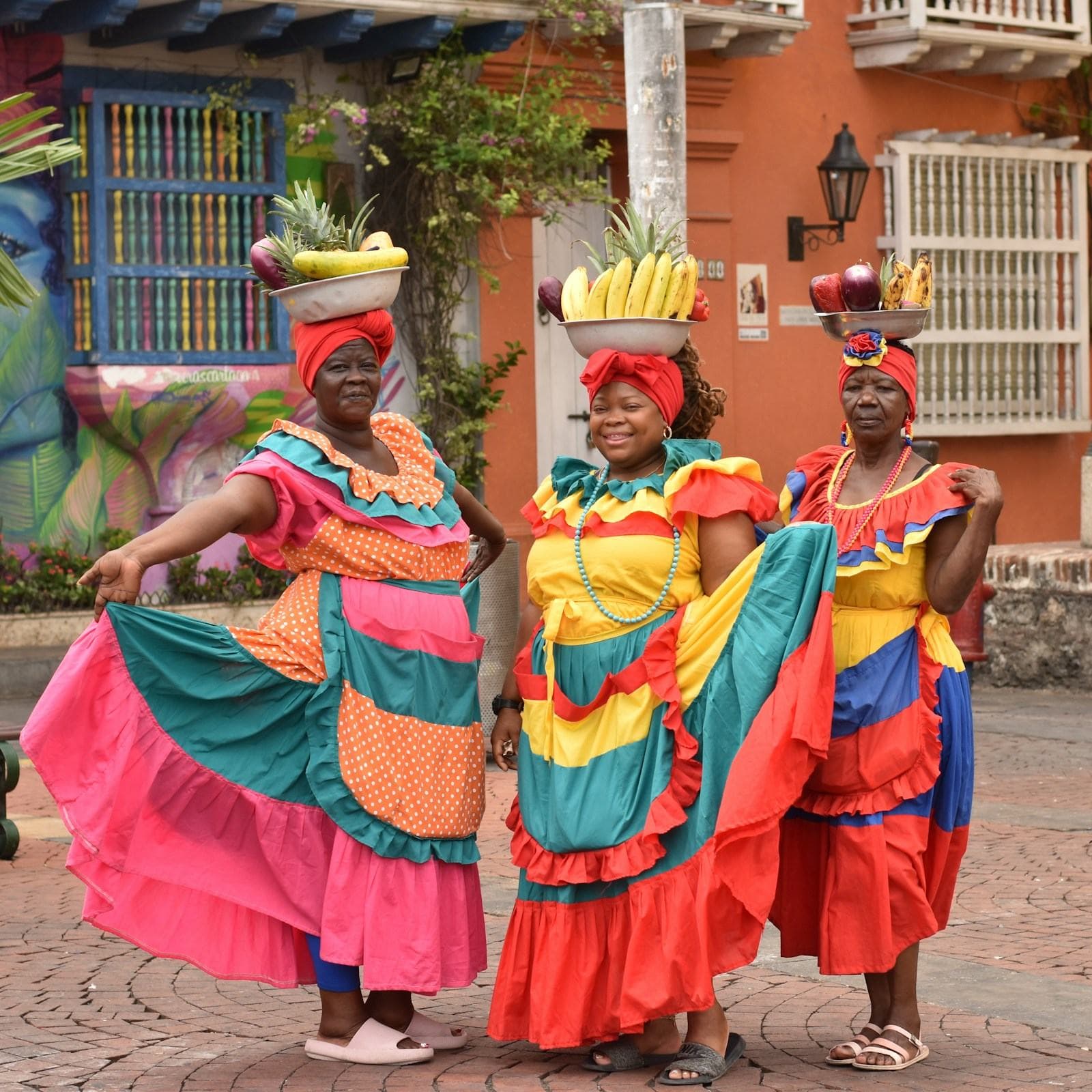 Three palenqueras in swirling pink, yellow and turquoise dresses balance fruit bowls in Cartagena