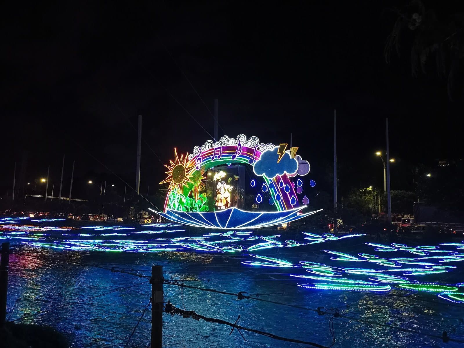 Glowing Christmas light sculptures floating on the Medellín River beside neon flower lights during Alumbrados