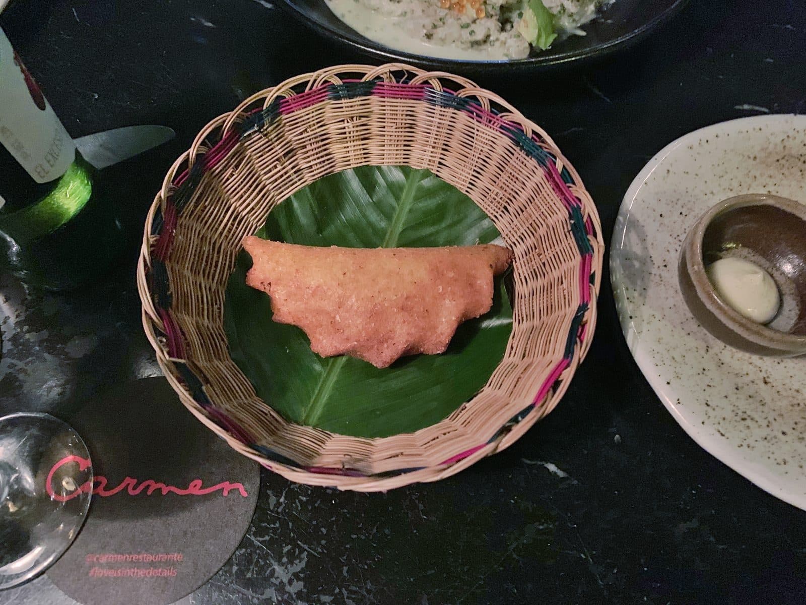 Golden empanada in a woven basket on a banana leaf beside Carmen's branded coaster