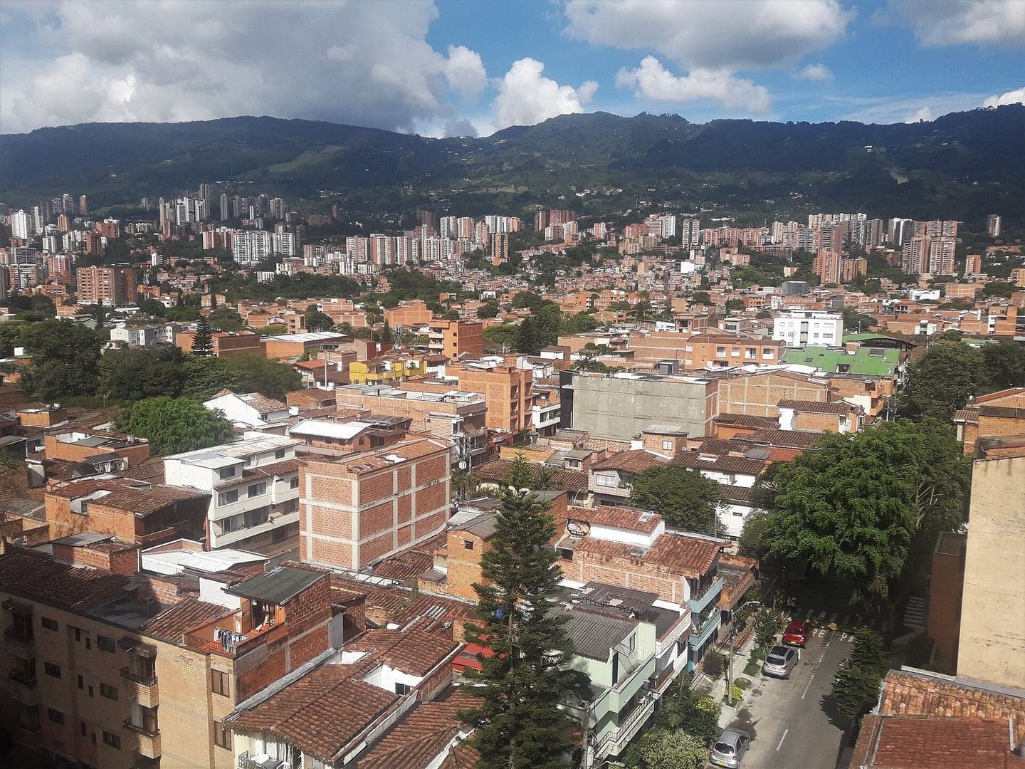 Red-brick rooftops of Envigado spreading toward green mountains under a bright partly cloudy sky