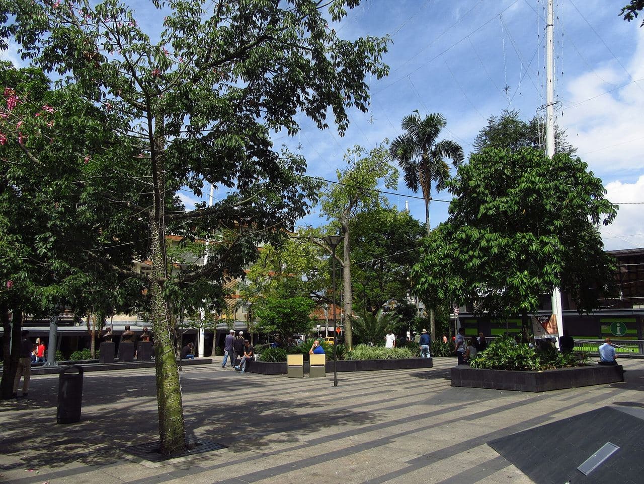 Shady trees and open plaza of Envigado's Parque Principal on a sunny afternoon