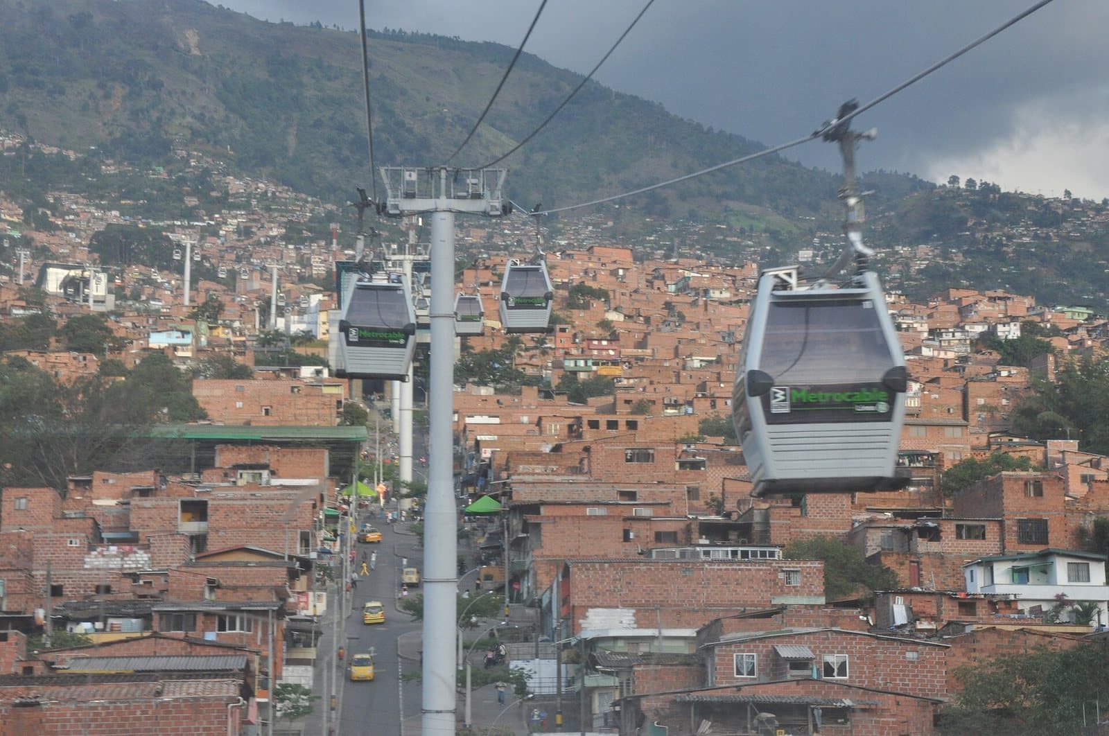 Metrocable gondolas climbing over red-brick hillside homes above a busy Medellín street