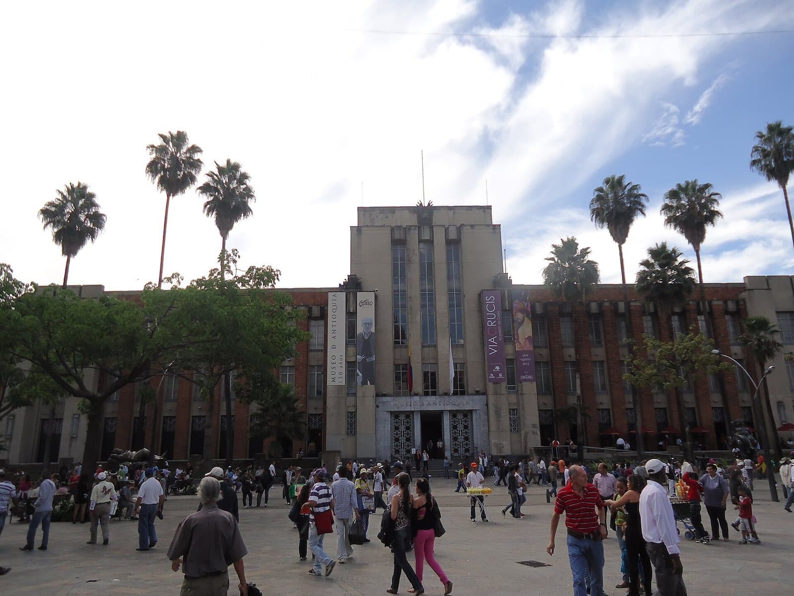 Art deco facade of the Museo de Antioquia with palm trees and visitors crossing Plaza Botero