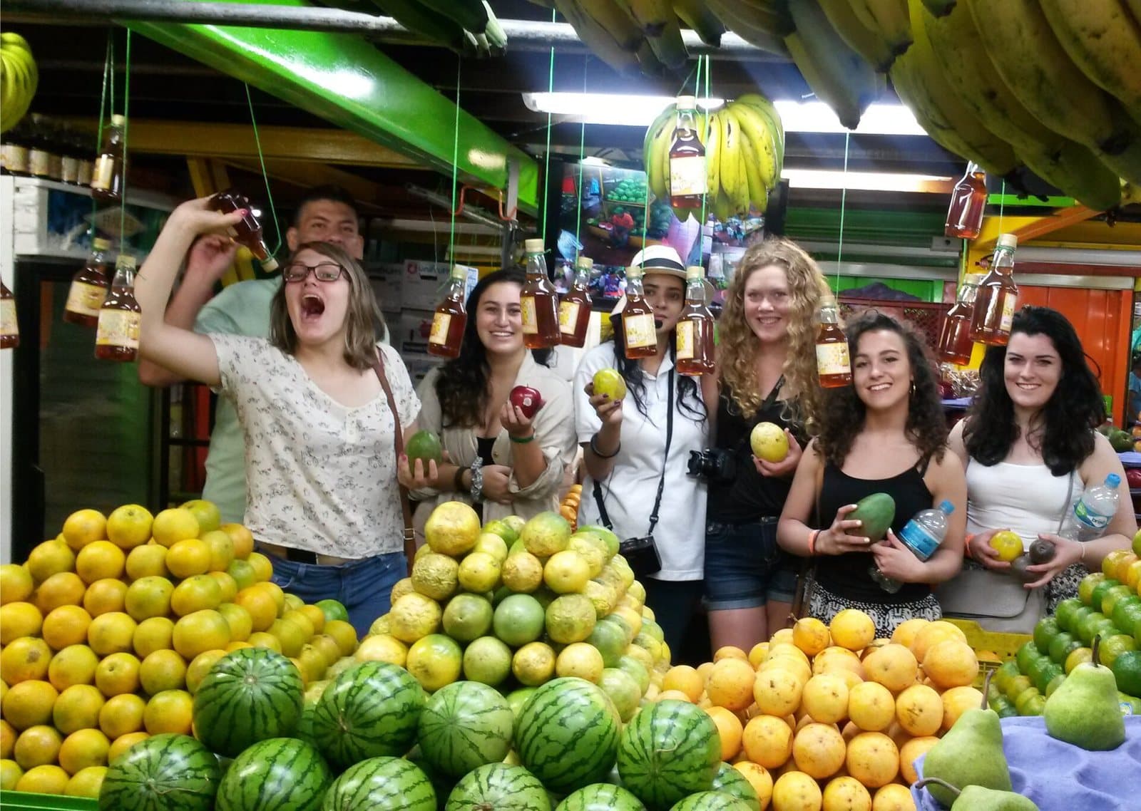 Visitantes sonrientes sosteniendo frutas exóticas detrás de montones de naranjas, sandías y mangos en la Plaza Minorista
