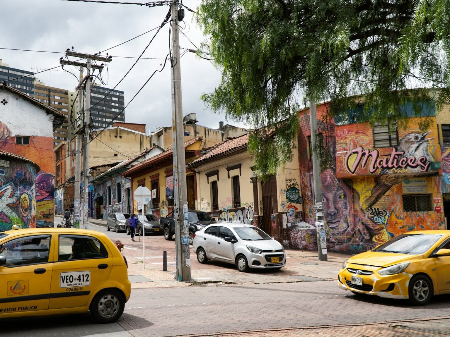Bright yellow taxis roll past mural-covered colonial houses on a sunny Bogotá street corner