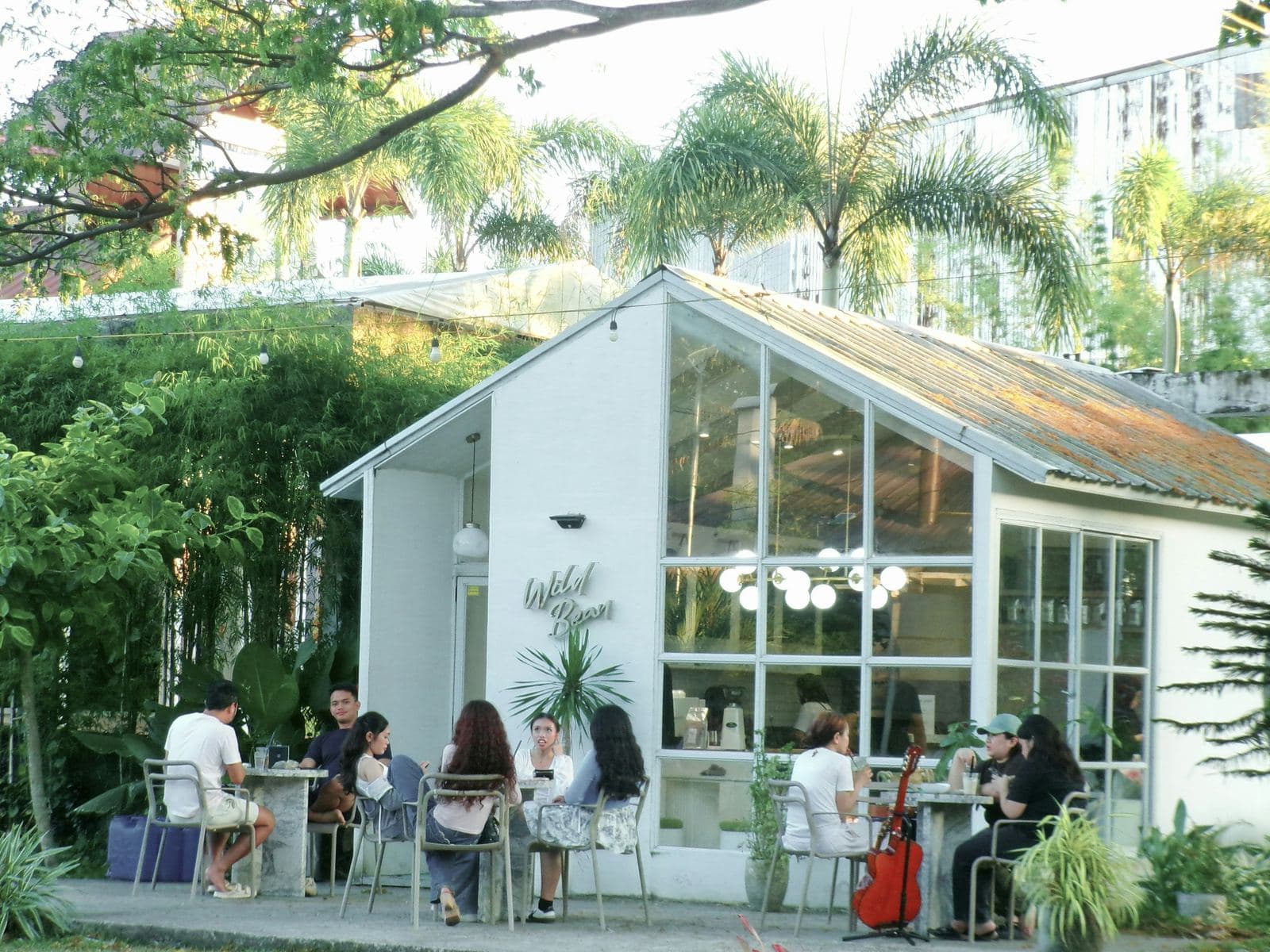 Friends chat at sunny tables outside a white glasshouse café surrounded by palms and plants
