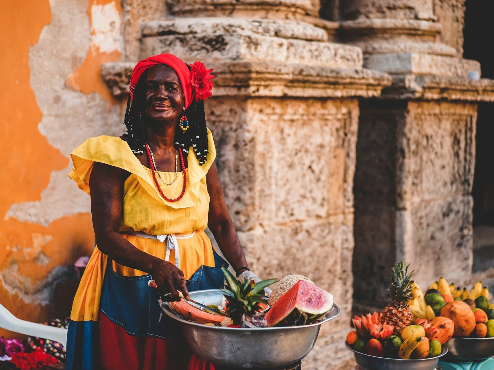 A smiling fruit vendor in a yellow dress and red headscarf offers fresh tropical fruit